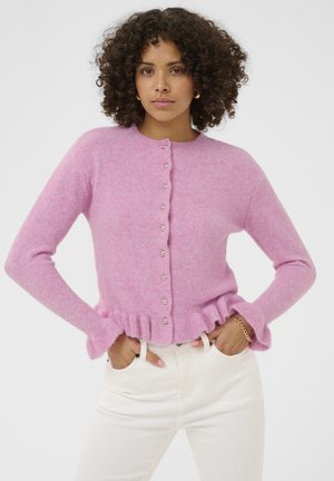 Kobieta z kręconymi włosami, ubrana w różowy sweter na guziki z falbaną u dołu i białe spodnie, z rękami w kieszeniach, na białym tle.