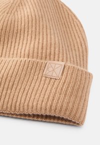 Beige knitted beanie with ribbed texture and a folded cuff. Features a small branded leather tag on the side. Soft wool material.