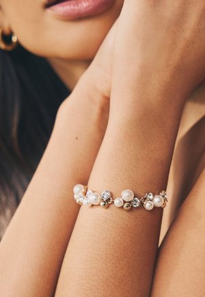 Woman wearing a gold bracelet adorned with pearls and clear crystals on her wrist, resting her arm near her face with gold hoop earrings.