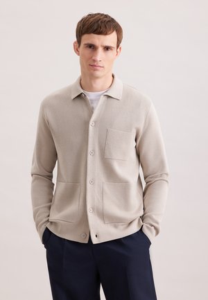 Jonge man met kort bruin haar, die een beige vest met knopen draagt over een wit shirt en donkere broek, staand met handen in de zakken.