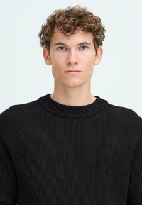 Svart ribbad tröja med rundad halsringning och raglanärmar, tillverkad av strukturerad stickad tyg, uppställd mot en enfärgad bakgrund.