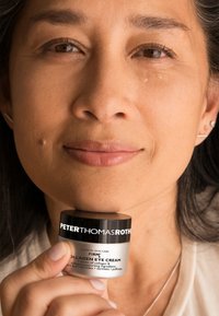 Peter Thomas Roth Firm Collagen Eye Cream i en svart och silverkruka som hålls nära ansiktet, med tydlig text på etiketten. Slät textur synlig.
