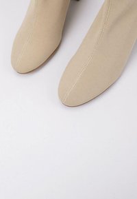 Botas de tobillo beige con parte superior de tejido texturizado, punta afilada y tacón delgado. La suela es marrón, lo que contrasta con el material superior.