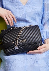 Czarna pikowana torebka na ramię z złotymi łańcuszkami, z logo "GUESS" i odpinaną metką. Miękka skórzana faktura, usztywniona konstrukcja.