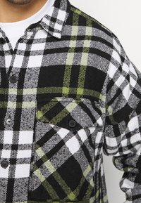 Camicia a scacchi bianca e nera con dettagli verdi, realizzata in tessuto morbido. Presenta un colletto, chiusura a bottoni e una tasca a toppa con bottoni.
