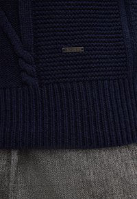 Dunkelblauer Strickpullover mit geripptem Saum und geflochtenem Detail über grauen Hosen im Fischgrätmuster. Metall-Label mit der Aufschrift "JOOP!".