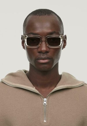Hanorac din polar cu fermoar, de culoare brown; ochelari de soare dreptunghiulari, cu lentile întunecate; textură netedă; design minimal. Fundal neutru.