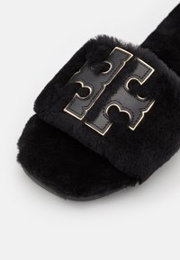 Chausson noir duveteux ouvert à l’avant avec une large bride comportant un emblème décoratif en cuir noir cousu et bordé d’or.