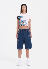 Camiseta rosa recortada con gráfico de dragón multicolor, combinada con pantalones cortos de mezclilla azules tipo bermuda que presentan detalles de flores en los bolsillos y bordados.