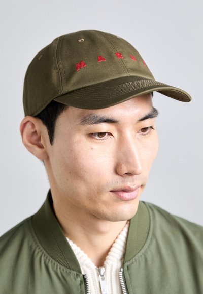 Gorra verde oliva con texto bordado en rojo, con ala curvada y diseño de seis paneles, llevada con una chaqueta bomber verde y un suéter crema.