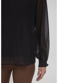 Sort plissert bluse med gennemsigtige lange ærmer, elastiske manchetter og en løs pasform. Stoffet har en glat tekstur og et strømmende design.