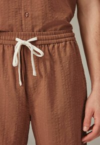 Jumpsuit marrone con texture, cinturino elastico e cordoncino bianco. Presenta un bottone nella parte superiore e un tessuto morbido e leggero.