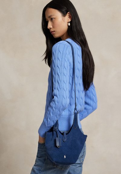 Blå mocka axelväska med kurvad form, sidoficka och justerbar rem. Modellen bär en kabelstickad tröja och jeans.