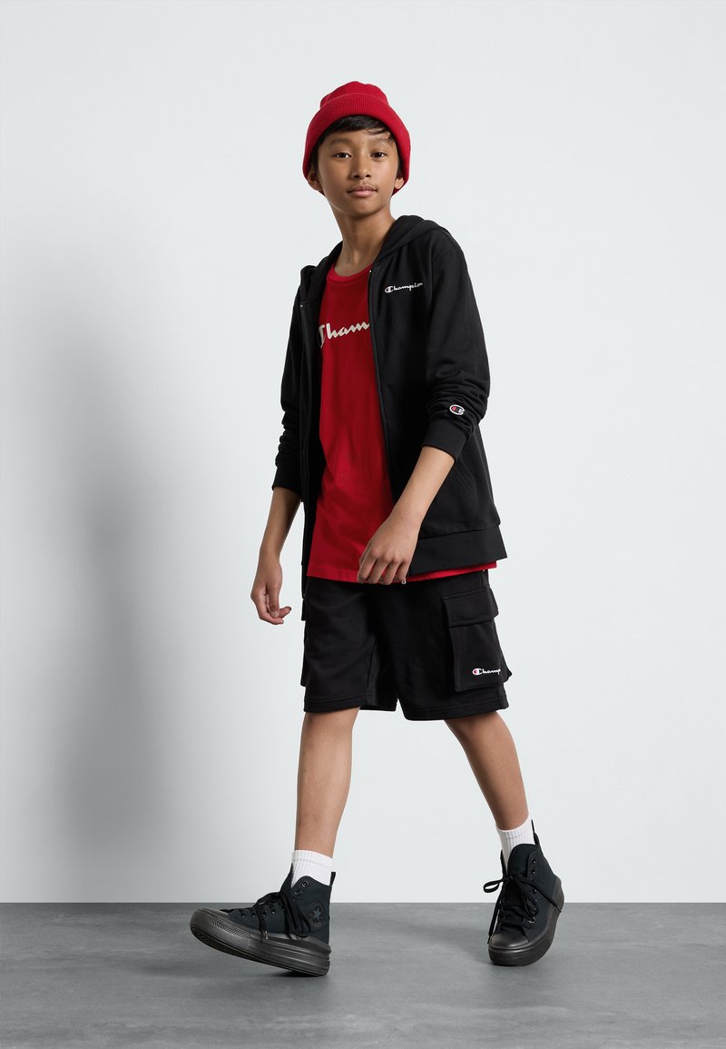Felpa nera con zip, t-shirt grafica rossa, pantaloni cargo neri, berretto rosso. Sneakers nere con dettagli bianchi. In piedi su un pavimento grigio, sfondo neutro.