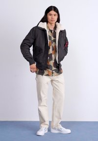 Jachetă bomber neagră cu guler din blană albă, cămașă cu model de camuflaj dedesubt, pantaloni beji deschiși �și adidași albi cu accente gri.