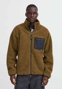Male model wearing brown textured fleece jacket with black chest pocket and zipper, standing against plain light background.