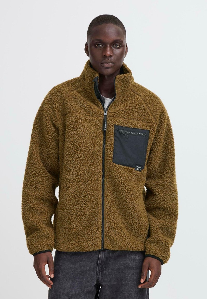 Male model wearing brown textured fleece jacket with black chest pocket and zipper, standing against plain light background.