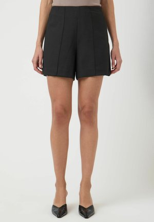 Kobieta ubrana w czarne, dopasowane szorty z wysokim stanem i czarne szpilki w szpic, stojąca z luźno opuszczonymi ramionami po bokach na jednolitym tle.