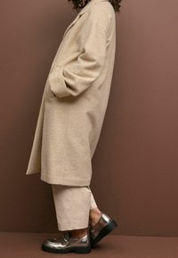Manteau beige texturé avec revers à cran et poches latérales, associé à un pantalon cropped beige clair et des chaussures métalliques argentées avec semelles noires.
