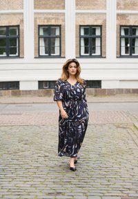 Vestido maxi con un patrón en espiral en azul y negro, escote en V y mangas cortas; combinado con zapatos de tacón negros sobre un pavimento de adoquines.