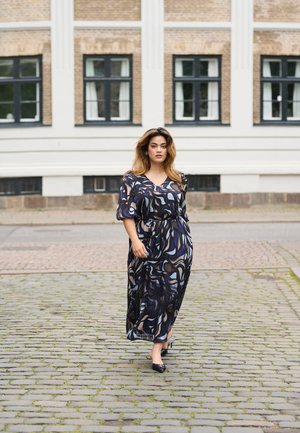 Robe maxi avec un motif tourbillonnant bleu et noir, encolure en V et manches courtes ; portée avec des chaussures à talons noires sur un pavé en pierre.