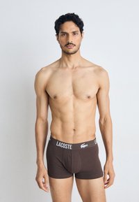 Rjave bombažne boksarice z črno-belo Lacoste logo na pasu, nosi moški model na nevtralni podlagi.