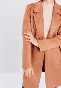 Ruskea mokkanahkainen takki, jossa on lovettu kaulus, suora leikkaus ja pitkät hihat. Takissa on etuaukko ja se on kerroksittain puettu kevyen tekstuurin neuleen päälle.
