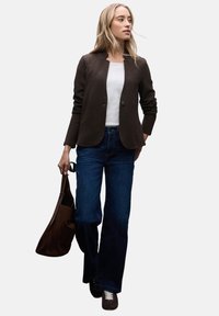 Hnědý texturovaný blazer přes bílý top, spárovan�ý s rozšířenými modrými džíny a tmavohnědými botami. Drží hnědou semišovou kabelku přes rameno.