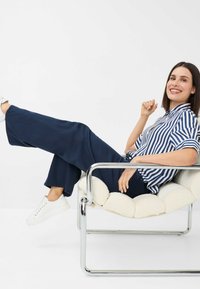 Striped blue and white shirt, navy wide-leg pants, and white sneakers. Sitting in a chrome-framed chair with a plush white seat cushion.