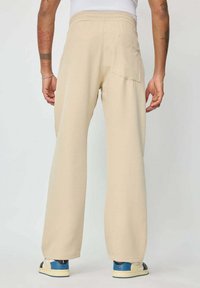 Pantalon large beige avec une ceinture élastique et une poche arrière. Le modèle porte des baskets bleu et noir, mettant en valeur une texture lisse.