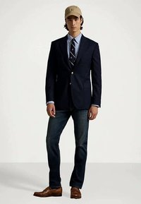 Άνδρας με καπέλο μπεζ, ναυτικό σακάκι, ριγέ γραβάτα, ανοιχτό μπλε πουκάμισο, σκούρα τζιν και καφέ δερμάτινα παπούτσια, στέκεται μπροστά σε ουδέτερο φόντο.