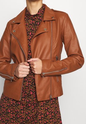 Osoba ubrana w brązową skórzaną kurtkę na czarnej sukience z pomarańczowymi i różowymi kwiatowymi wzorami, ręce trzymają przednie krawędzie kurtki.