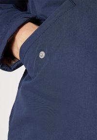 Tissu de veste bleu marine avec des coutures visibles, présentant un accent de bouton-pression métallique sur une poche latérale. Texture lisse sans motifs.