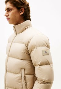 Beżowa kurtka puchowa z wysokim kołnierzem, pikowanym wzorem i naszywką marki na lewym rękawie. Posiada zamek błyskawiczny i boczne kieszenie.