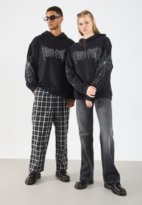 Svarte hettegensere med spindelvev-grafikk; den mannlige modellen har rutete bukser, og den kvinnelige modellen har grå jeans. Begge har svarte sko.