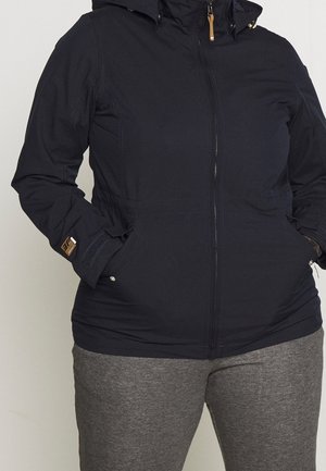 Persoon draagt een zwarte jas met ritssluiting en capuchon, handen in de zakken, gecombineerd met grijze broek, tegen een effen lichte achtergrond.