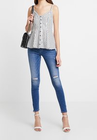 Top peplum sin tirantes a rayas en blanco y negro con botones en la parte delantera, combinado con jeans skinny azules desgastados y tacones blancos con tiras al tobillo. Bolso negro.