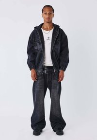 Zwarte spijkerjasje met elastische manchetten, gecombineerd met loszittende zwarte spijkerbroeken. De outfit bevat een wit T-shirt met logo. Zwarte sneakers.