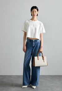 Maglietta corta bianca con scritte in rilievo, jeans a gamba larga blu e una borsa crema con dettagli marroni. Scarpe bianche a punta completano il look.