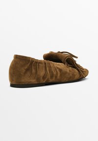 Brown suede loafers with a soft, textured surface, featuring a fringe detail and gathered sides, flat sole, and a rounded toe design.