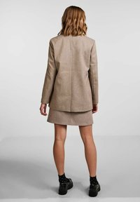 Kobieta stojąca tyłem, ubrana w beżową marynarkę i pasującą spódnicę, z czarnymi skarpetkami w kropki oraz czarnymi butami na jednolitym tle.