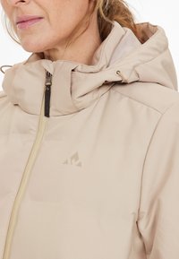 Beige vetoketjullinen takki hupulla, joka on pehmeää materiaalia, hienovarainen logo detail ja vaaleanvärinen vetoketju. Monikerroksinen kankaan suunnittelu.