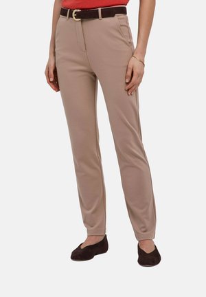 Vrouw die beige rechte broek draagt met een zwarte riem, rood topje en zwarte platte schoenen, staand tegen een egale achtergrond.