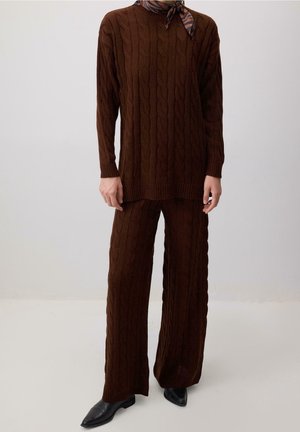 Osoba ubrana w brązowy sweter w warkocze i pasujące szerokie spodnie, czarne spiczaste buty oraz wzorzysty szalik na szyi.