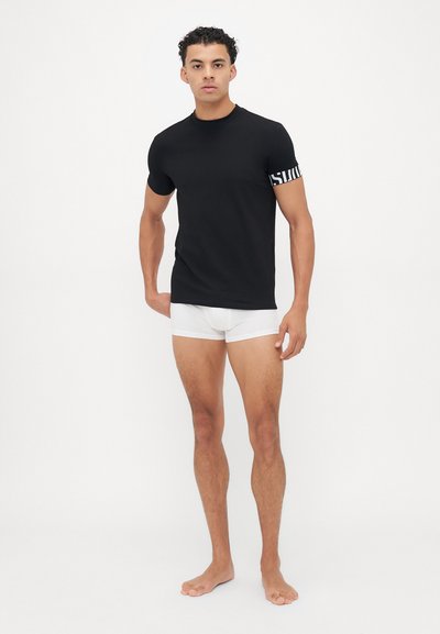 Schwarzes Kurzarm-T-Shirt mit Rundhalsausschnitt und weißen grafischen Akzenten an den Ärmeln, kombiniert mit weißen Boxershorts und barfuß.