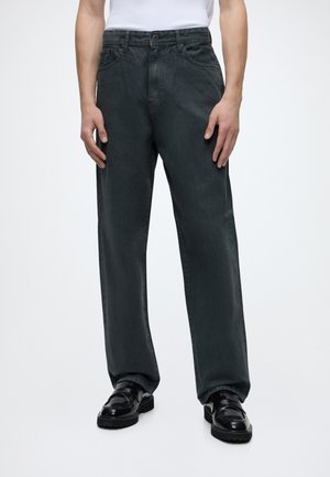 Homme portant un jean noir droit, une chemise blanche et des chaussures en cuir noires, debout devant un fond uni.