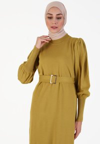 Vestido de suéter mostaza con cuello redondo, mangas abullonadas y cintura ceñida con una hebilla rectangular dorada. Textura suave.