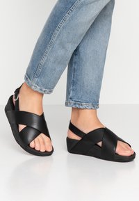 Fötter i svarta läder sandaletter med korsande remmar, ihop med ljusblå avklippta jeans mot en neutral bakgrund.