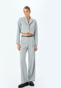Blazer recortado gris claro con un solo botón y mangas largas, combinado con pantalones de pierna ancha de cintura alta y zapatos de cuero negros.