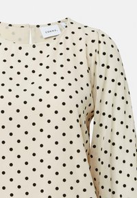 Cremige Bluse mit schwarzen Punkten, rundem Ausschnitt und gepufften Ärmeln, aus leichtem Stoff mit glatter Textur.